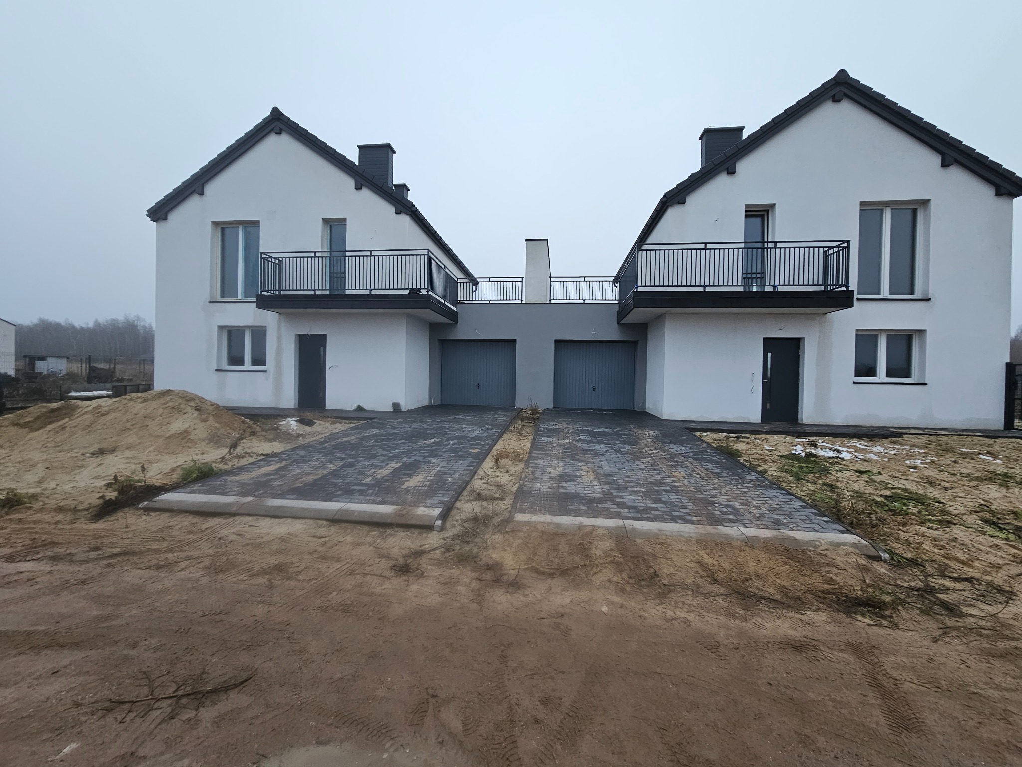 Dwa identyczne domy bliźniacze z czarnymi balustradami balkonowymi i szarą łączącą je konstrukcją nad garażami, na tle pochmurnego nieba.