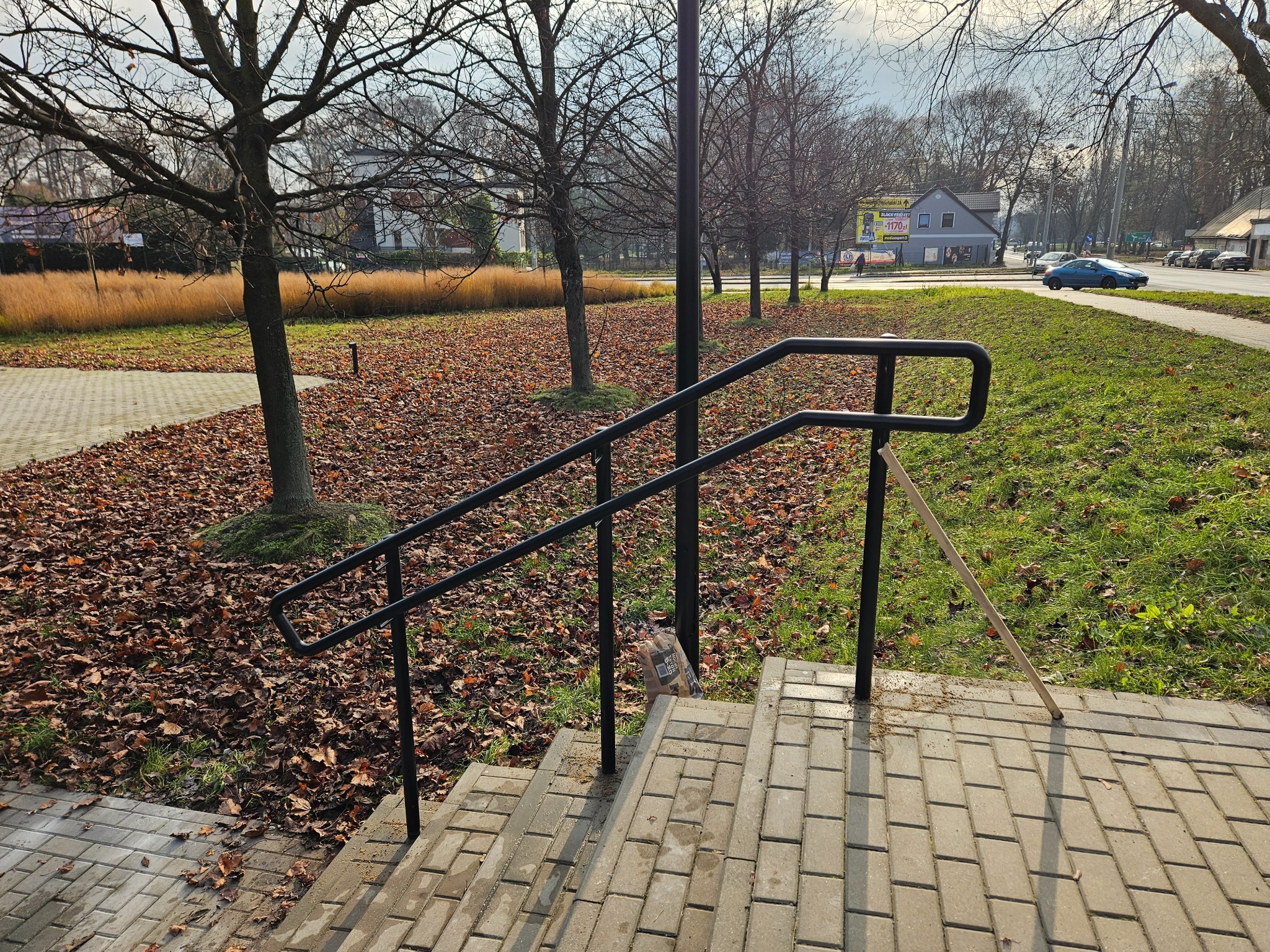 Czarna, metalowa poręcz schodowa na zewnątrz, z widocznymi śladami montażu na kostce brukowej. Tło stanowią drzewa i opadłe liście, co sugeruje jesienną porę roku.