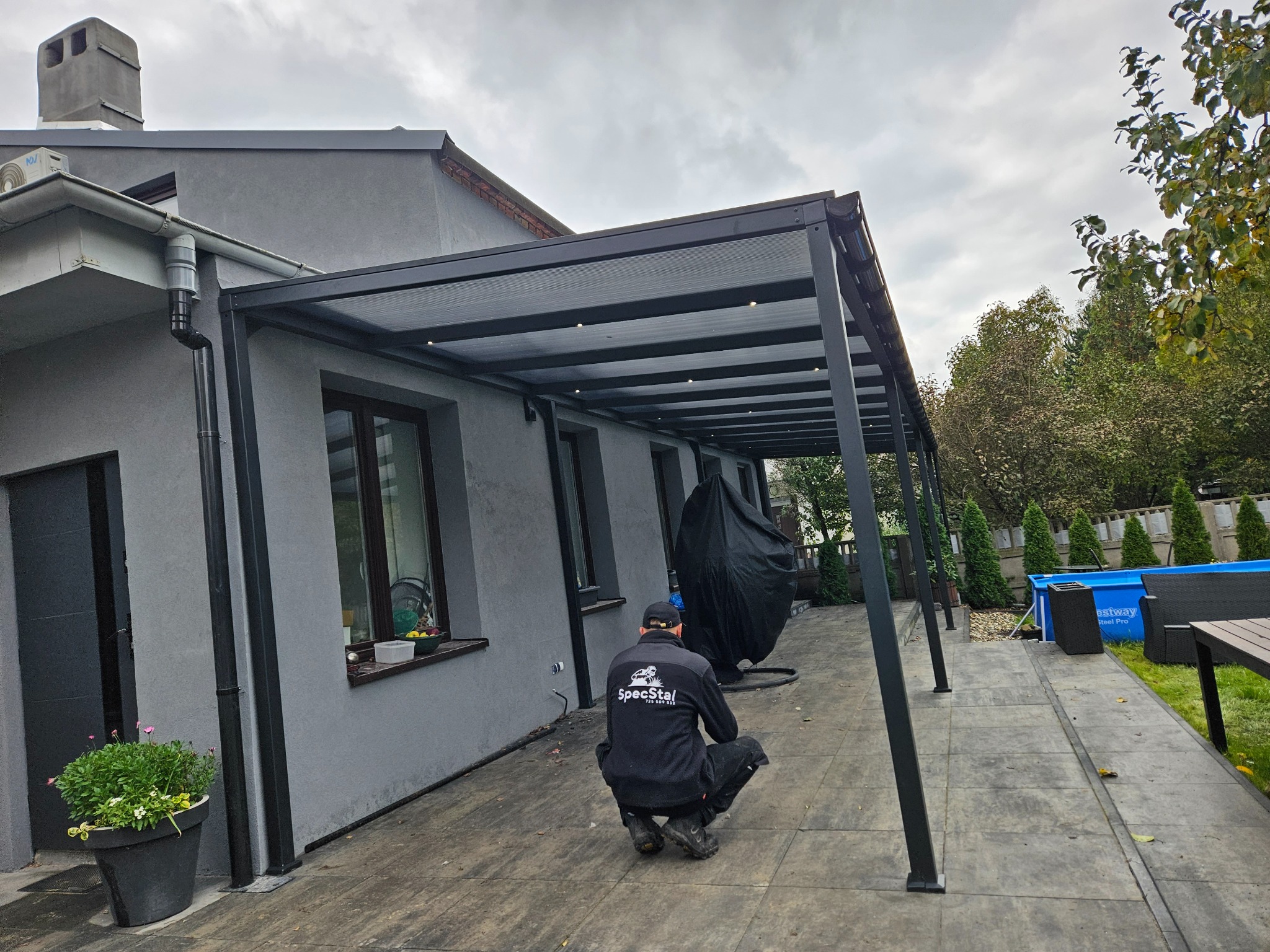 Nowoczesna, grafitowa pergola z oświetleniem LED, przylegająca do domu. Pracownik firmy instalacyjnej w trakcie pracy. W tle basen i zieleń ogrodu.