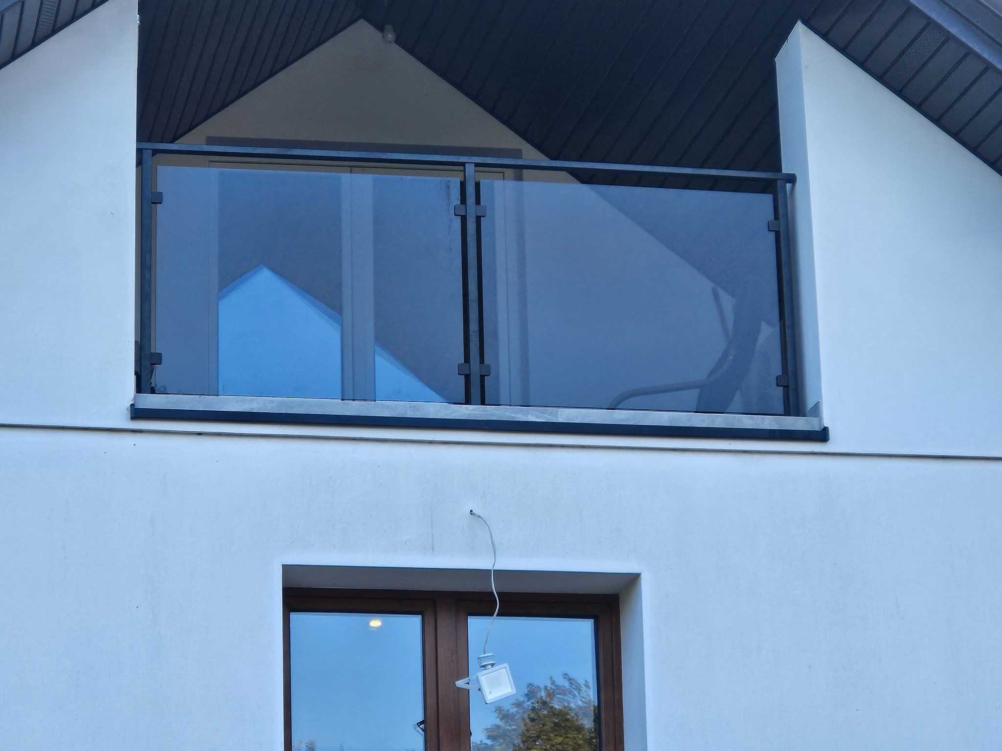 Nowoczesna balustrada szklana z czarnymi profilami na balkonie domu z białymi ścianami. Widoczny fragment okna i lampa zewnętrzna. Minimalistyczny design.