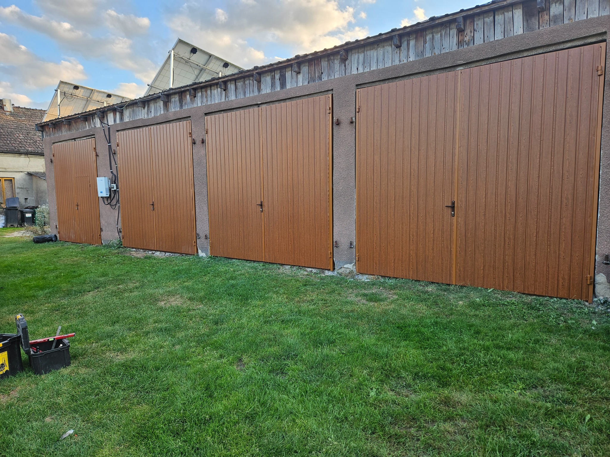 Brązowe bramy garażowe z pionowymi panelami, zamontowane w rzędzie w budynku z panelami słonecznymi na dachu. Na pierwszym planie narzędzia na trawie.