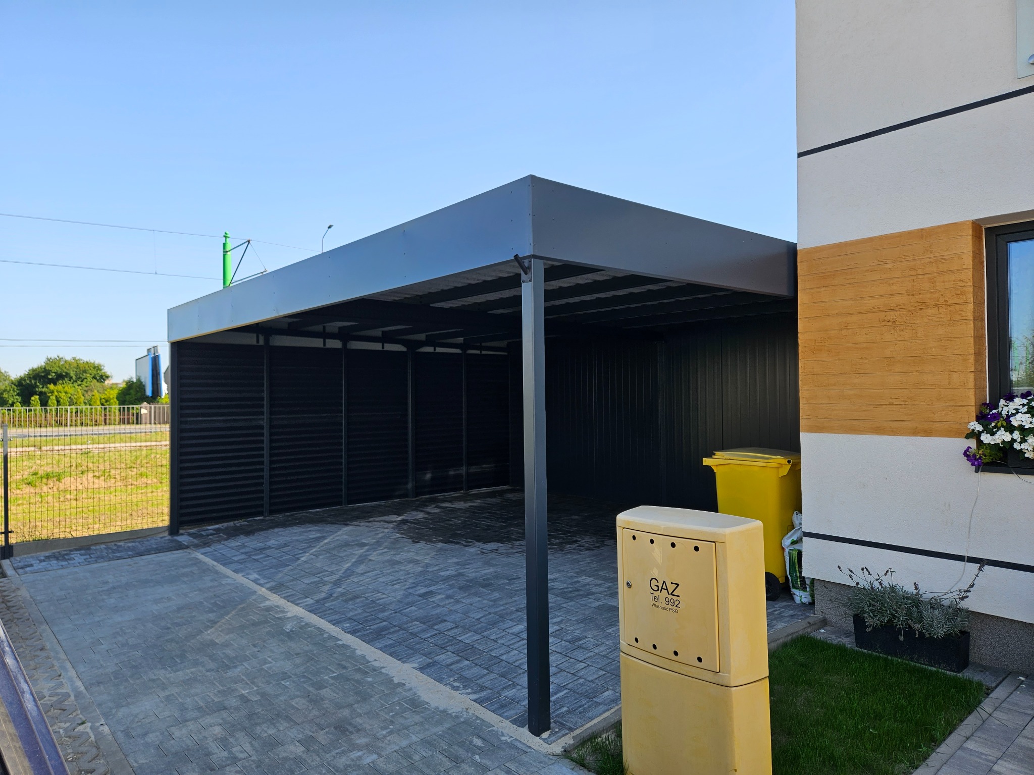 Nowoczesna, czarna wiata garażowa z metalowymi panelami, przylegająca do budynku z żółtym licznikiem gazu i koszem na śmieci. Kostka brukowa na podjeździe.