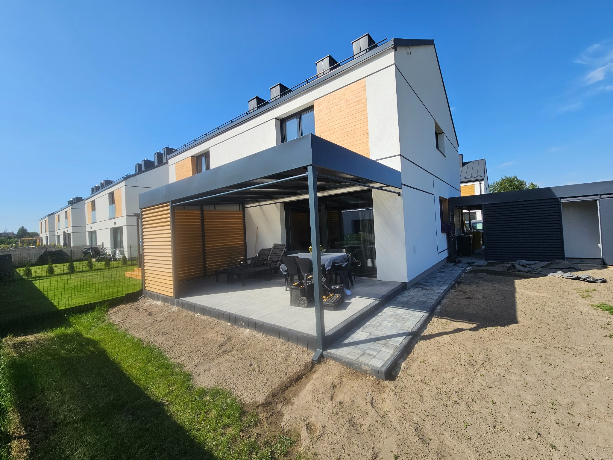 Nowoczesna pergola z metalową konstrukcją i drewnianymi lamelami, przylegająca do budynku w zabudowie szeregowej. Widoczne meble ogrodowe i kostka brukowa.