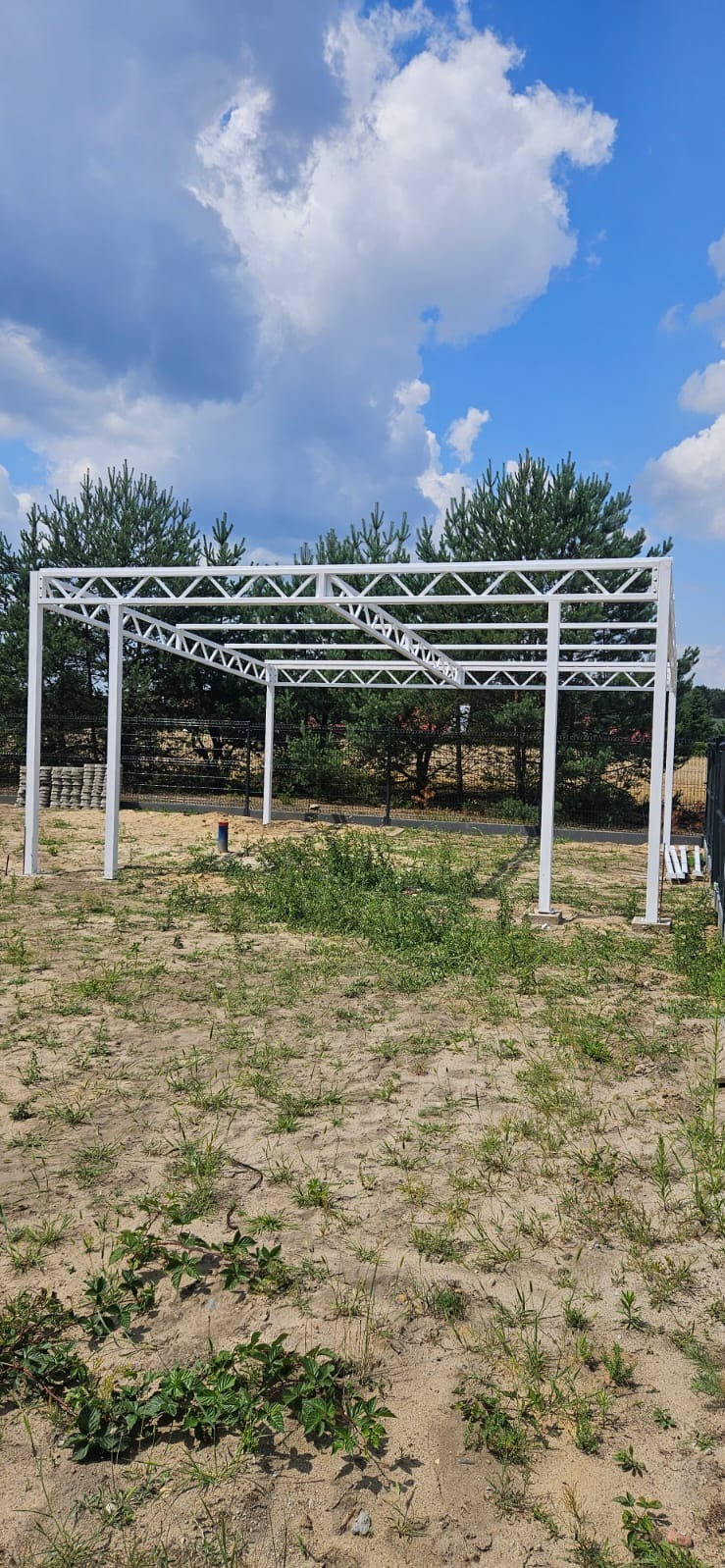 Biała, metalowa konstrukcja pergoli na tle błękitnego nieba z chmurami i zieleni drzew. Widoczne elementy spawane. Konstrukcja w surowym stanie, bez zadaszenia.