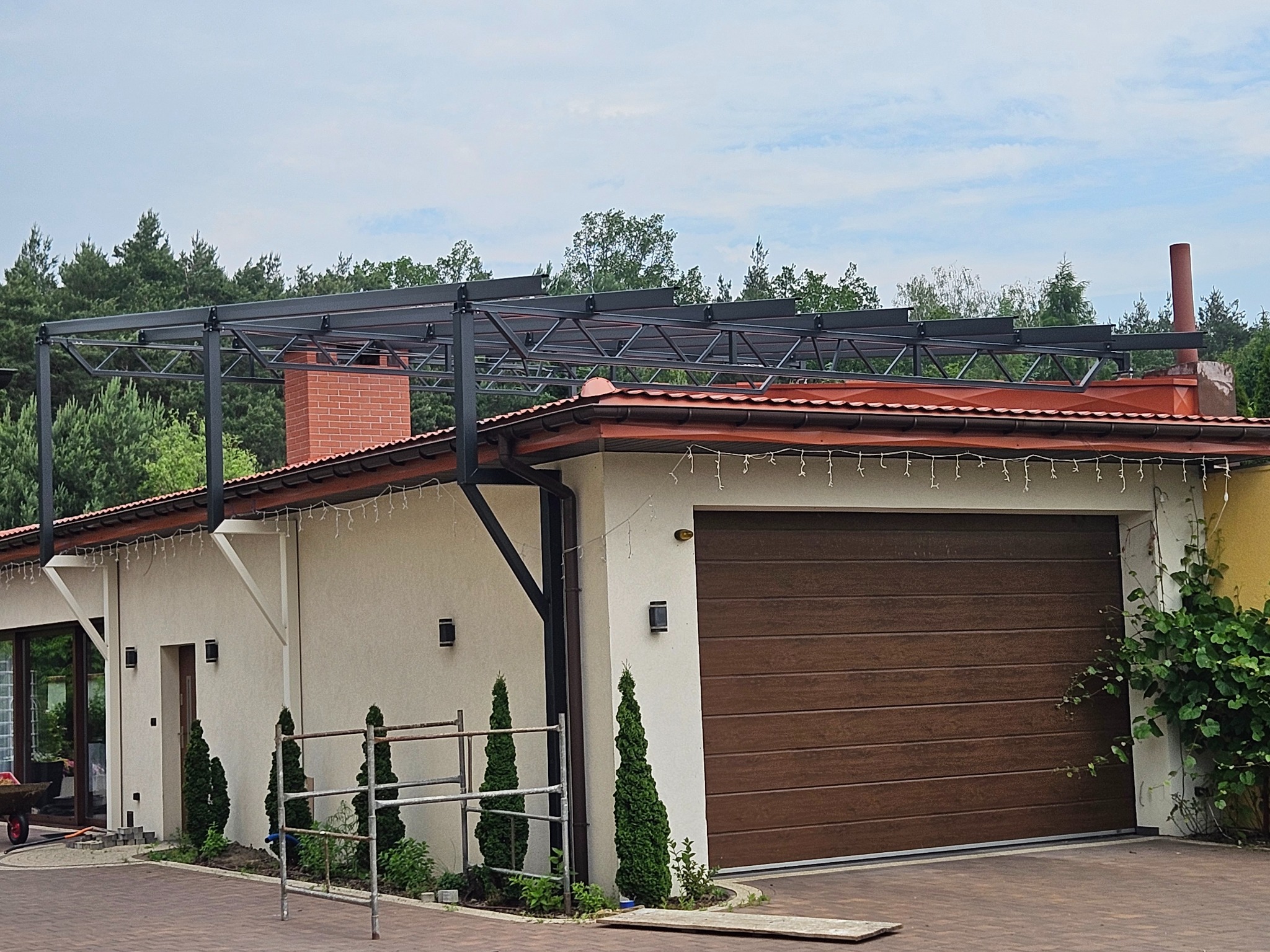 Czarna konstrukcja metalowa pergoli nad budynkiem z brązową bramą garażową i ceglanym kominem. W tle drzewa i niebo. Widoczne elementy montażowe.