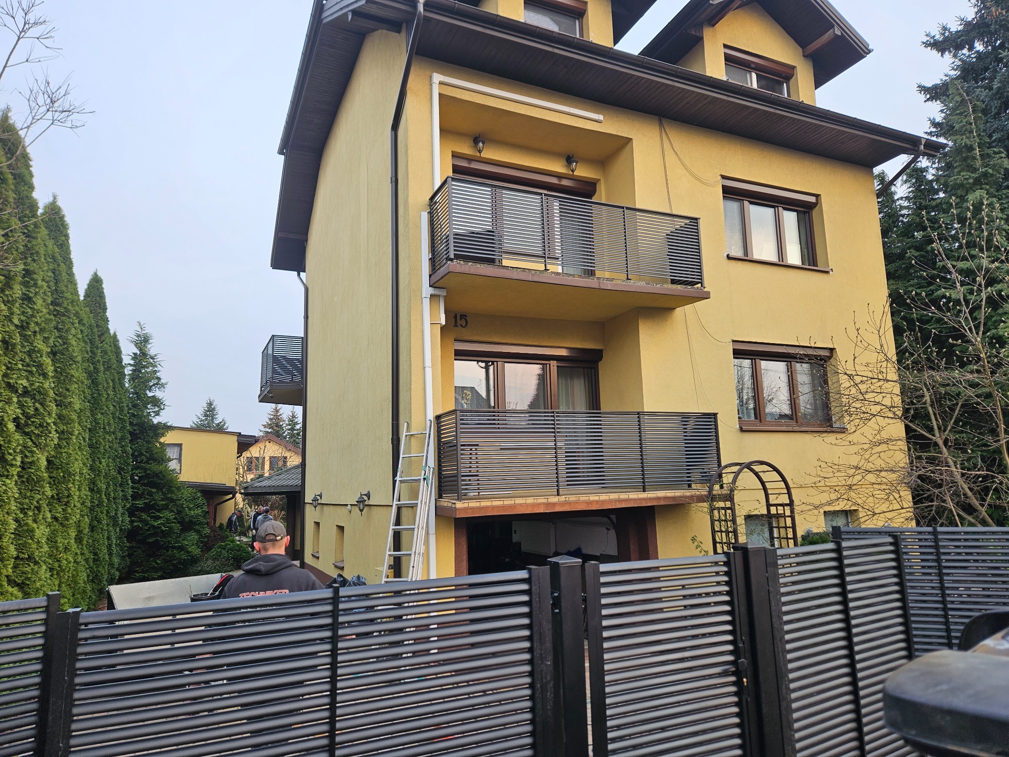 Żółty dom z balkonami zabezpieczonymi poziomymi, metalowymi panelami. W tle drabina oparta o balkon. Na pierwszym planie fragment ogrodzenia z paneli.
