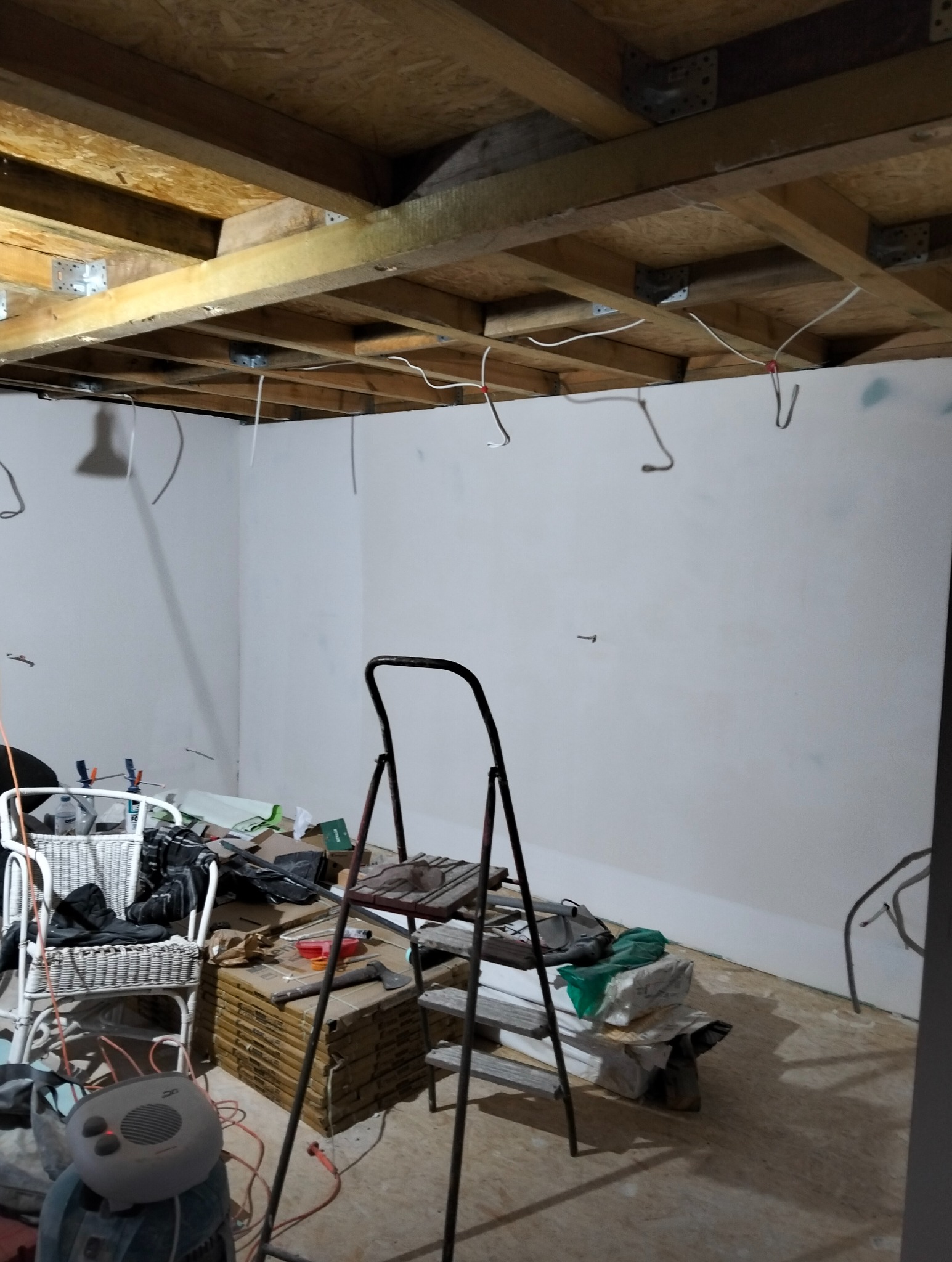 Wnętrze w trakcie remontu: białe ściany, drewniany strop z widocznymi kablami, drabina, materiały budowlane i narzędzia. Etap przygotowania do dalszych prac wykończeniowych.