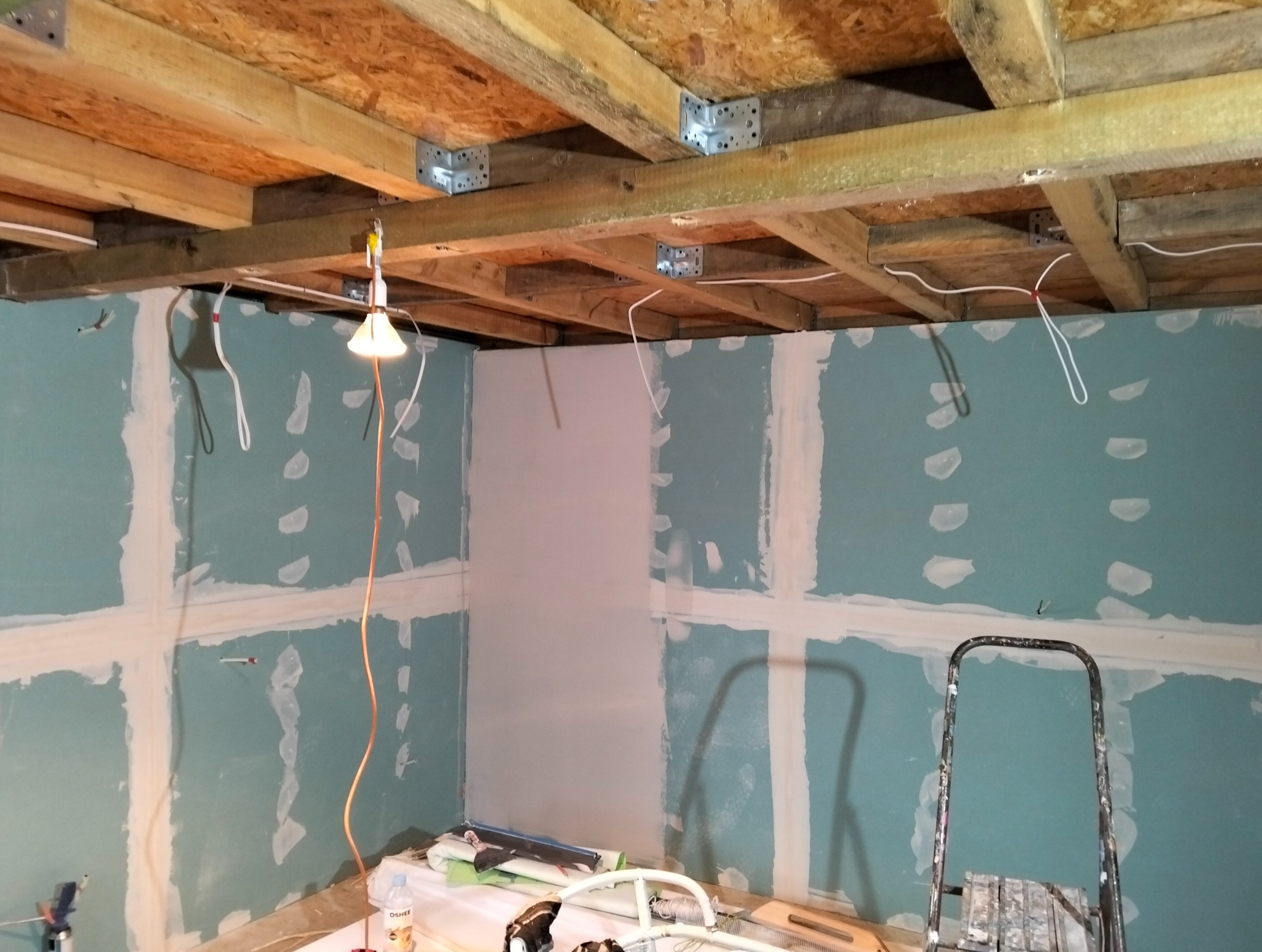 Ściana z płyt gipsowo-kartonowych w trakcie szpachlowania, widoczne łączenia i fragment gładzi. Na suficie drewniana konstrukcja z elementami metalowymi.