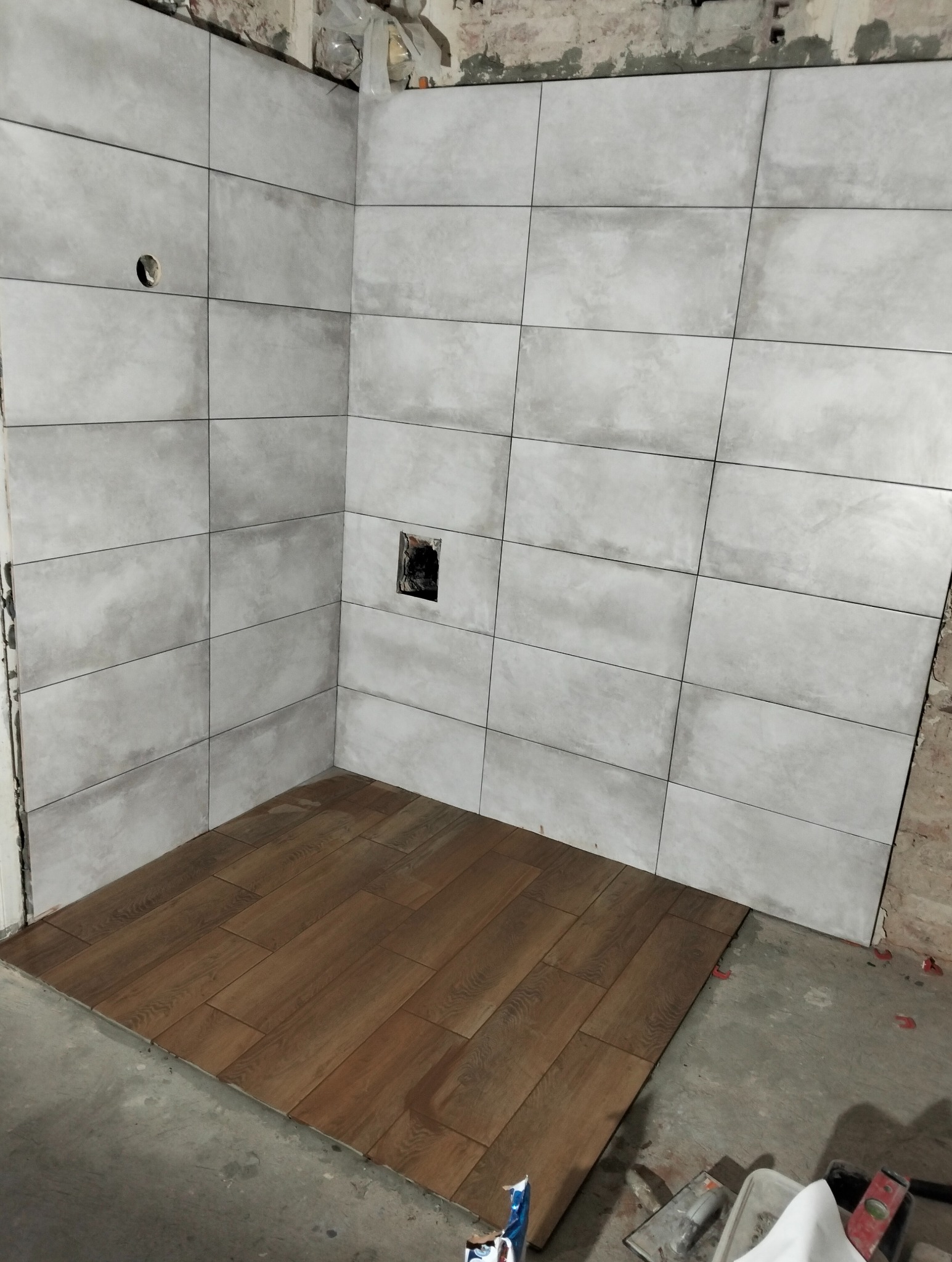 Wykończenie łazienki: szare, matowe płytki ścienne ułożone w regularny wzór, kontrastujące z drewnopodobną podłogą. Widoczne narzędzia i surowe ściany w tle.