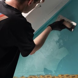 ZUBRA SPÓŁKA Z OGRANICZONĄ ODPOWIEDZIALNOŚCIĄ - Mężczyzna w czarnej koszulce i rękawicach szpachluje turkusową ścianę. Widoczne taśmy malarskie przy podłodze i suficie. Używa pacy do rozprowadzania gładzi.