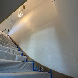 ZUBRA SPÓŁKA Z OGRANICZONĄ ODPOWIEDZIALNOŚCIĄ - Ściana po szpachlowaniu dekoracyjnym w klatce schodowej, zabezpieczone schody folią i taśmą malarską. Widoczny fragment lampy i instalacji elektrycznej.