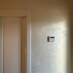 ZUBRA SPÓŁKA Z OGRANICZONĄ ODPOWIEDZIALNOŚCIĄ - Ściana z dekoracyjnym szpachlowaniem w odcieniach beżu, obok drzwi przesuwnych i panelu sterowania. Subtelne oświetlenie podkreśla fakturę.