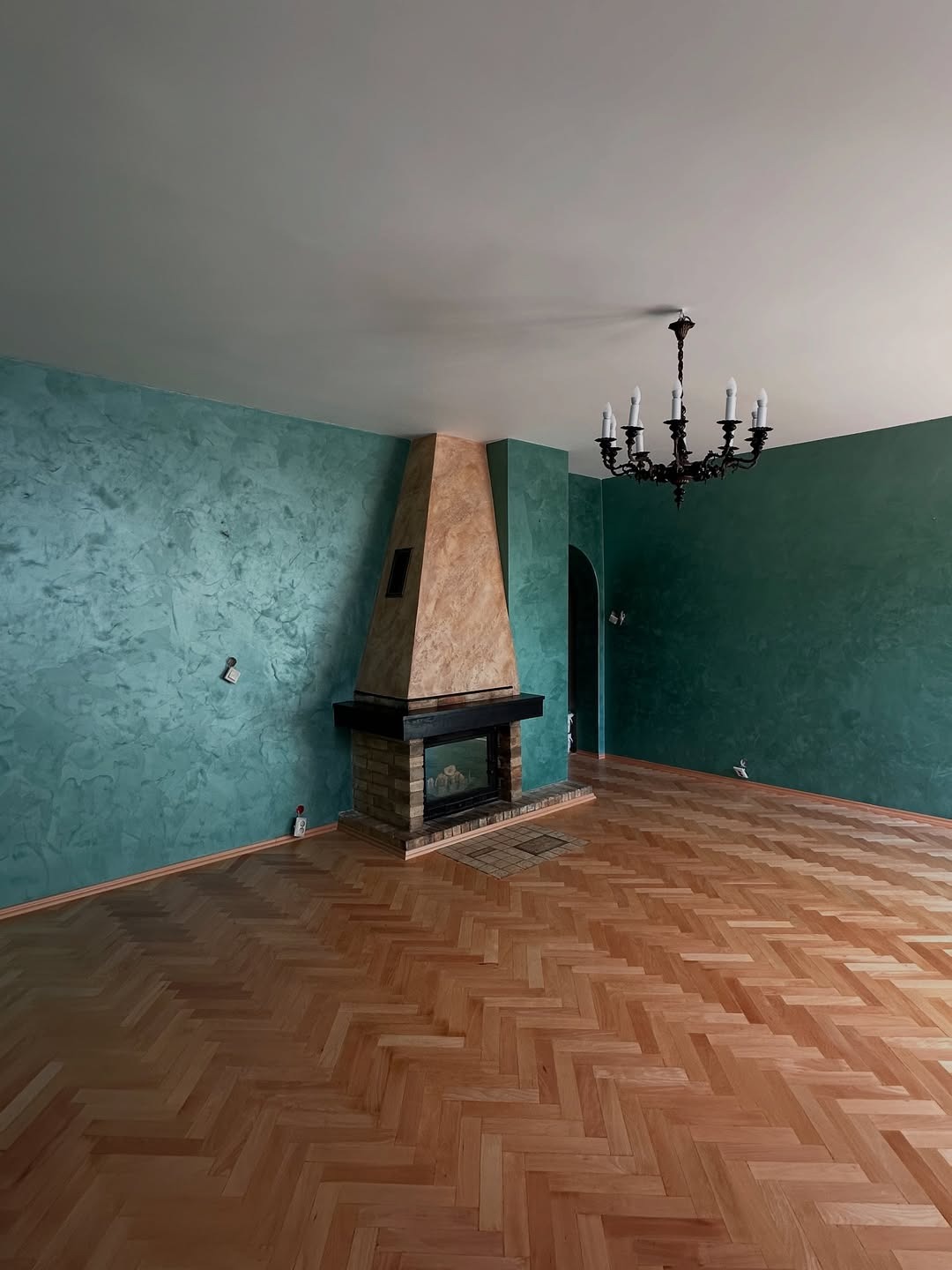 Salon po malowaniu ścian na turkusowo i zielono z efektem dekoracyjnym, parkiet w jodełkę, kominek z cegły i beżową górą, żyrandol.