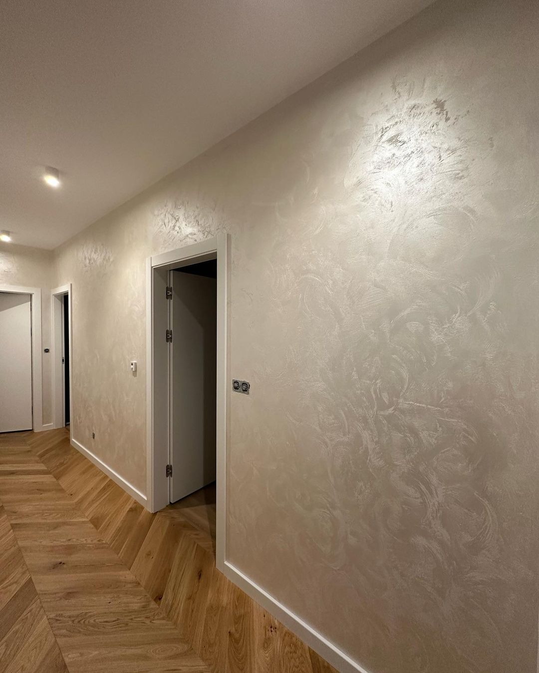 Korytarz z parkietem w jodełkę i ścianami pokrytymi dekoracyjnym, perłowym tynkiem. Widoczne białe listwy przypodłogowe i drzwi.