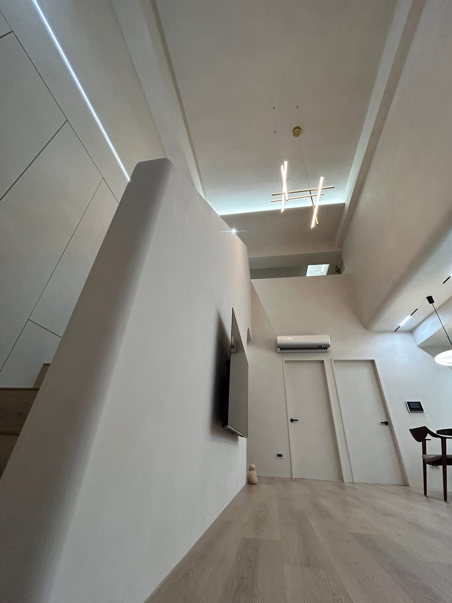 Minimalistyczne wnętrze z białymi ścianami, drewnianą podłogą i nowoczesnym oświetleniem. Widoczny fragment schodów, telewizor i drzwi. Charakterystyczny design.