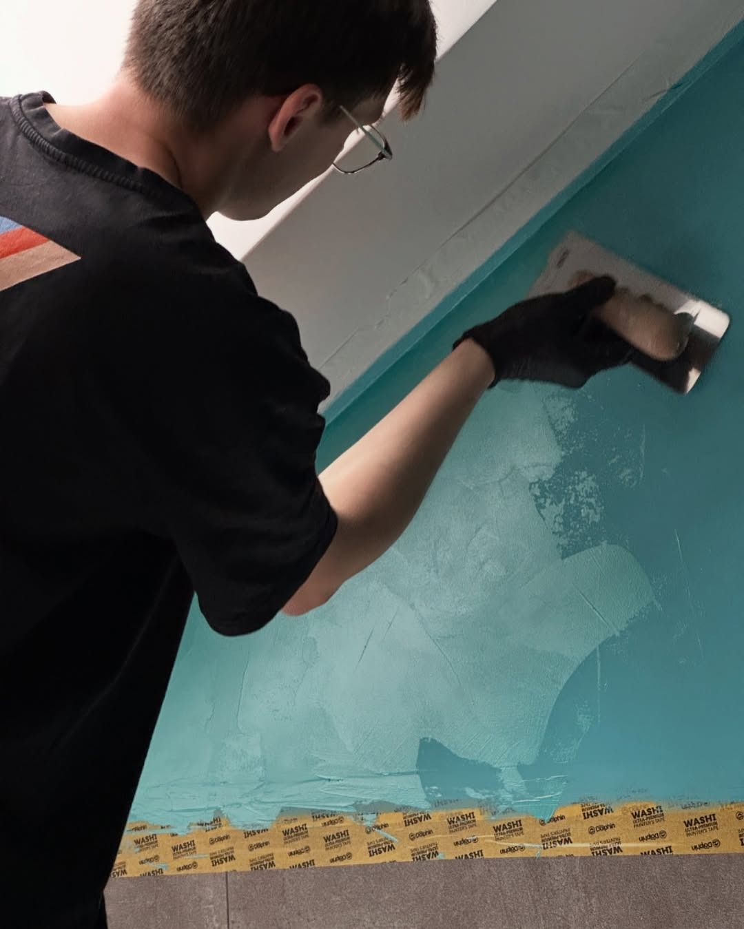 Mężczyzna w czarnej koszulce i rękawicach szpachluje turkusową ścianę. Widoczne taśmy malarskie przy podłodze i suficie. Używa pacy do rozprowadzania gładzi.