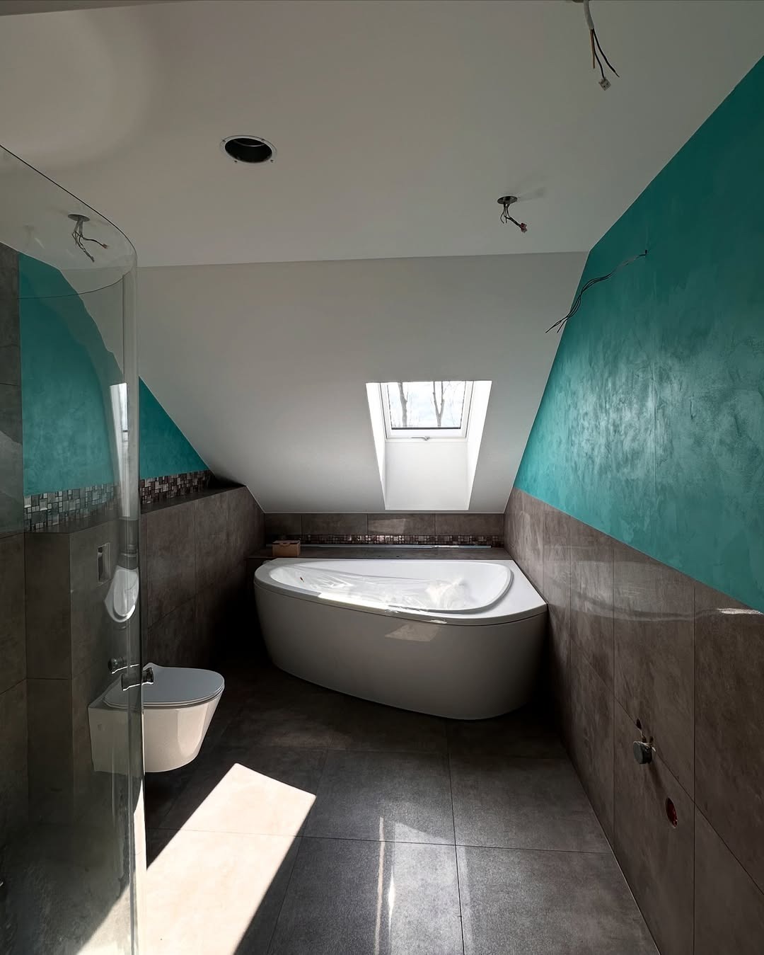 Nowoczesna łazienka na poddaszu z wolnostojącą wanną, szarą glazurą i turkusową ścianą. Widoczne niedokończone elementy elektryczne.