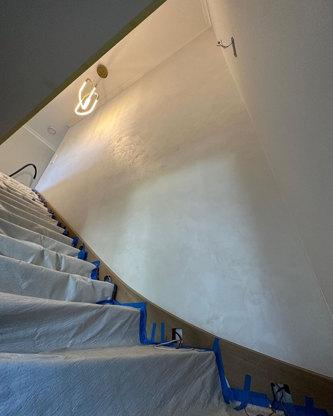 Ściana po szpachlowaniu dekoracyjnym w klatce schodowej, zabezpieczone schody folią i taśmą malarską. Widoczny fragment lampy i instalacji elektrycznej.