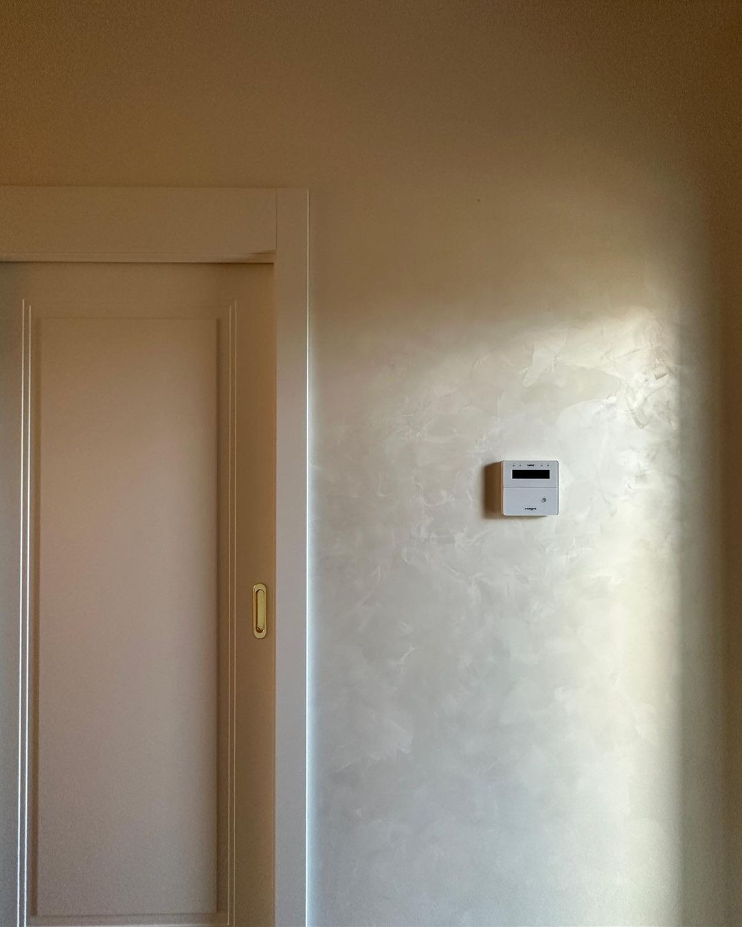 Ściana z dekoracyjnym szpachlowaniem w odcieniach beżu, obok drzwi przesuwnych i panelu sterowania. Subtelne oświetlenie podkreśla fakturę.