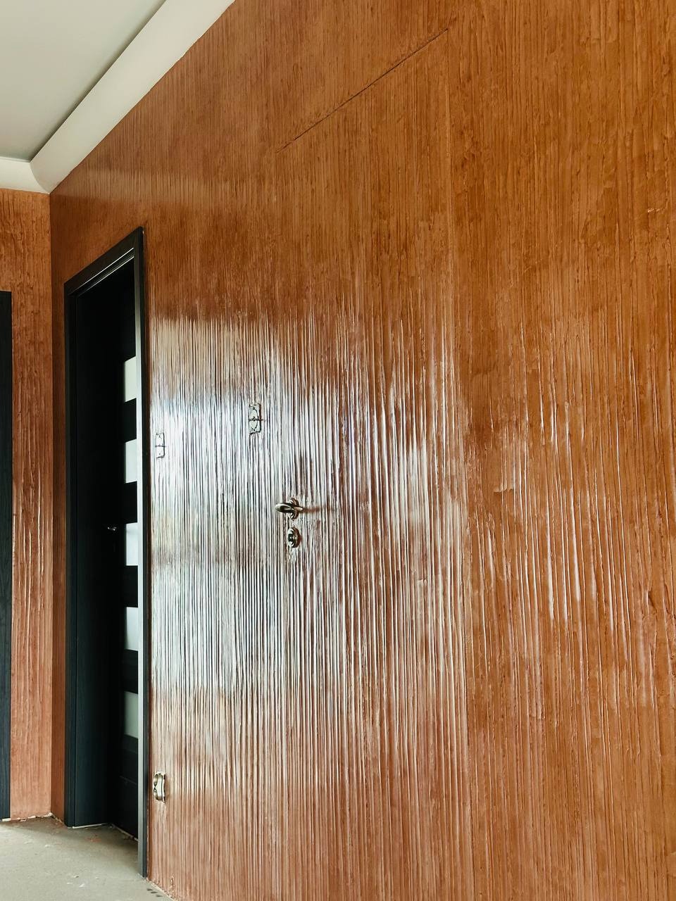 Teksturowana ściana w odcieniach brązu z widocznymi pionowymi żłobieniami, drzwi wejściowe z czarną ościeżnicą i srebrną klamką. Wykończenie wnętrza.