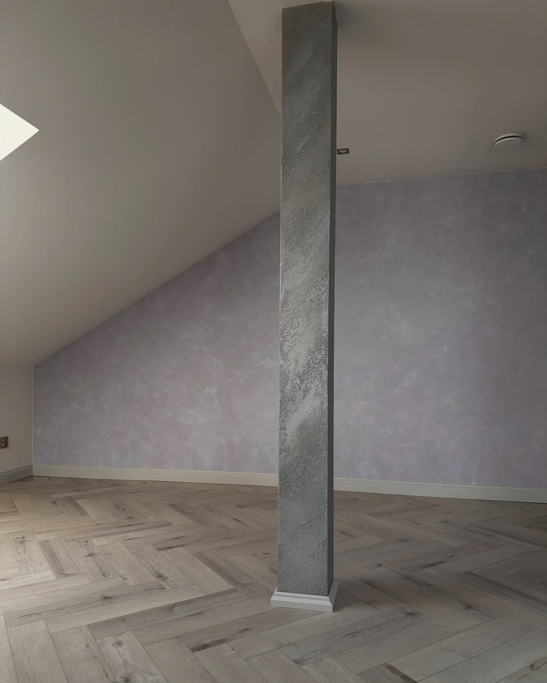 Pokój na poddaszu po malowaniu ścian z efektem dekoracyjnym, drewniana podłoga w jodełkę, kolumna w szarym kolorze. Widok na skośny sufit i okno dachowe.