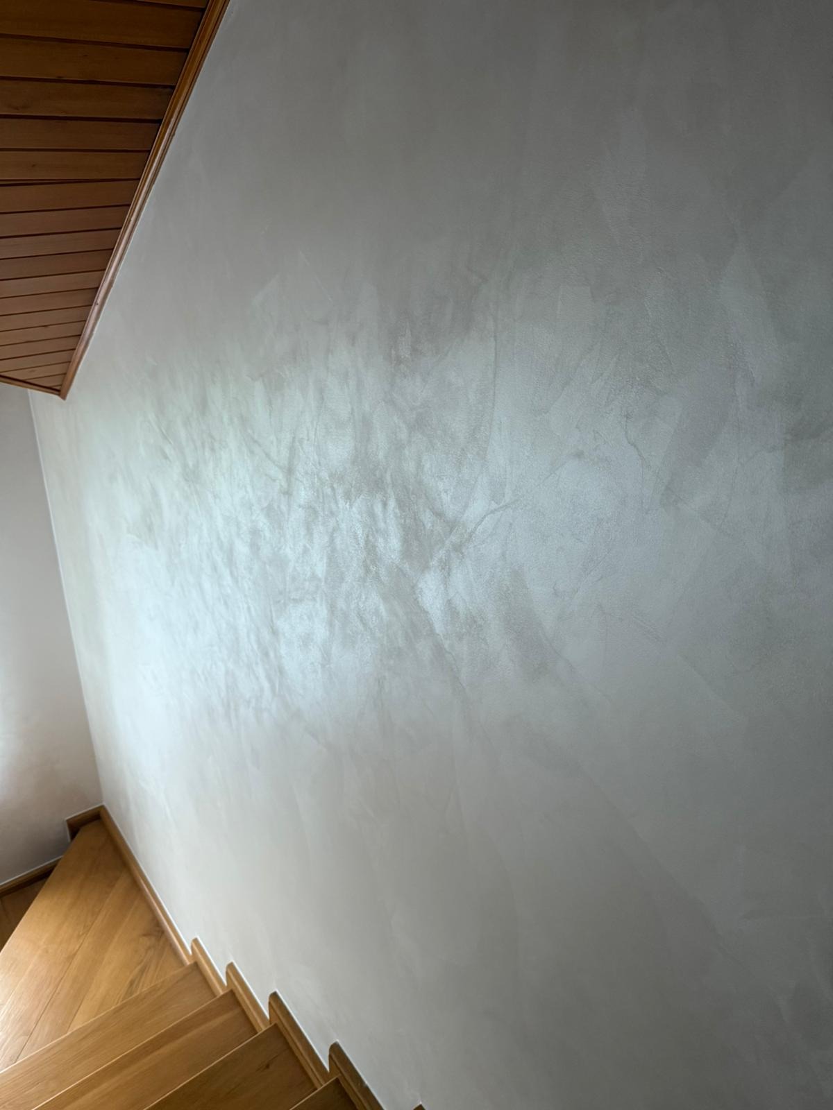 Ściana z efektem dekoracyjnym w odcieniach szarości, widoczna z perspektywy schodów z drewnianymi stopniami i boazerią sufitową. Subtelne refleksy światła podkreślają fakturę.