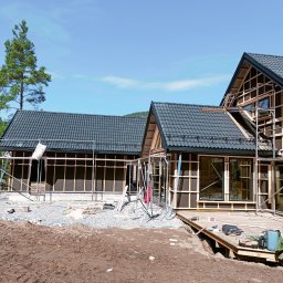 Marcin Okrasiński - Budowa domu z czarnym dachem w trakcie realizacji. Widoczne rusztowania, drewniana konstrukcja i częściowo ułożona dachówka. W tle zielone drzewo i błękitne niebo.