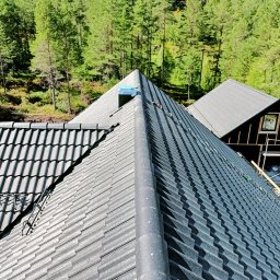 Marcin Okrasiński - Szary dach z dachówki ceramicznej, widok z góry na tle zielonego lasu. Na dachu widoczne elementy montażowe i komin w trakcie instalacji. Obok budynek w stanie surowym.