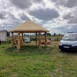 Marcin Okrasiński - Surowa drewniana altana z bali na trawiastym terenie, częściowo ukończona, z widocznym stosem drewna i samochodem dostawczym w tle, pod pochmurnym niebem.