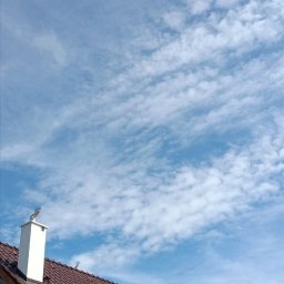 Marcin Okrasiński - Brązowy dach z dachówkami i oknami połaciowymi na tle błękitnego nieba z chmurami. Widoczny komin z ozdobną figurką. Budynek w trakcie budowy.