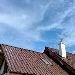 Marcin Okrasiński - Nowy, brązowy dach z dachówki ceramicznej, z oknem dachowym i rynną, na budynku w trakcie budowy. W tle błękitne niebo z chmurami.