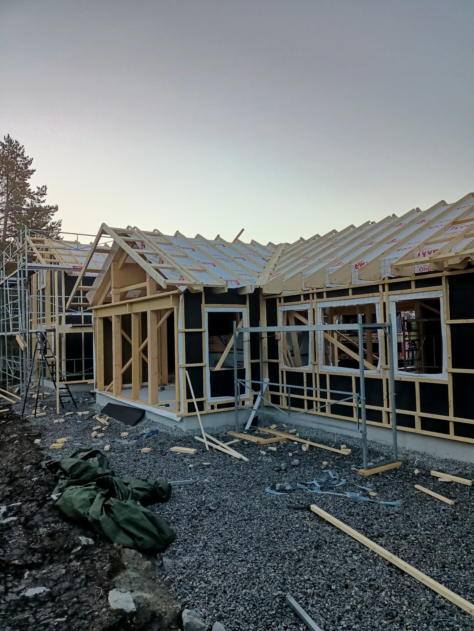 Budowa domu jednorodzinnego w stanie surowym otwartym. Widoczna drewniana konstrukcja ścian i dachu, czarna membrana, rusztowania i materiały budowlane na żwirowym podłożu.