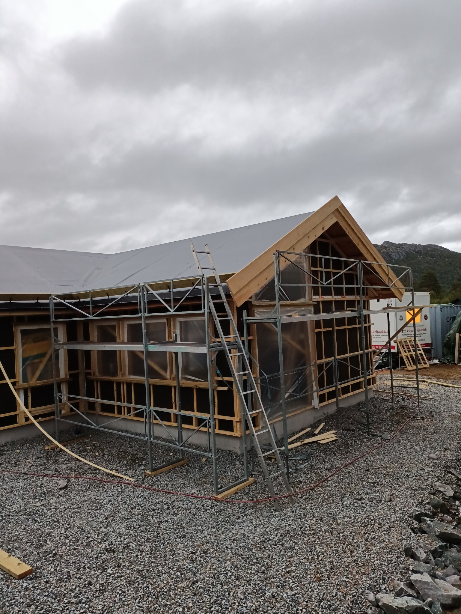 Wznoszenie konstrukcji dachu w Maszewie. Widoczna drewniana więźba, rusztowania i zabezpieczenie folią. W tle góry i pochmurne niebo.