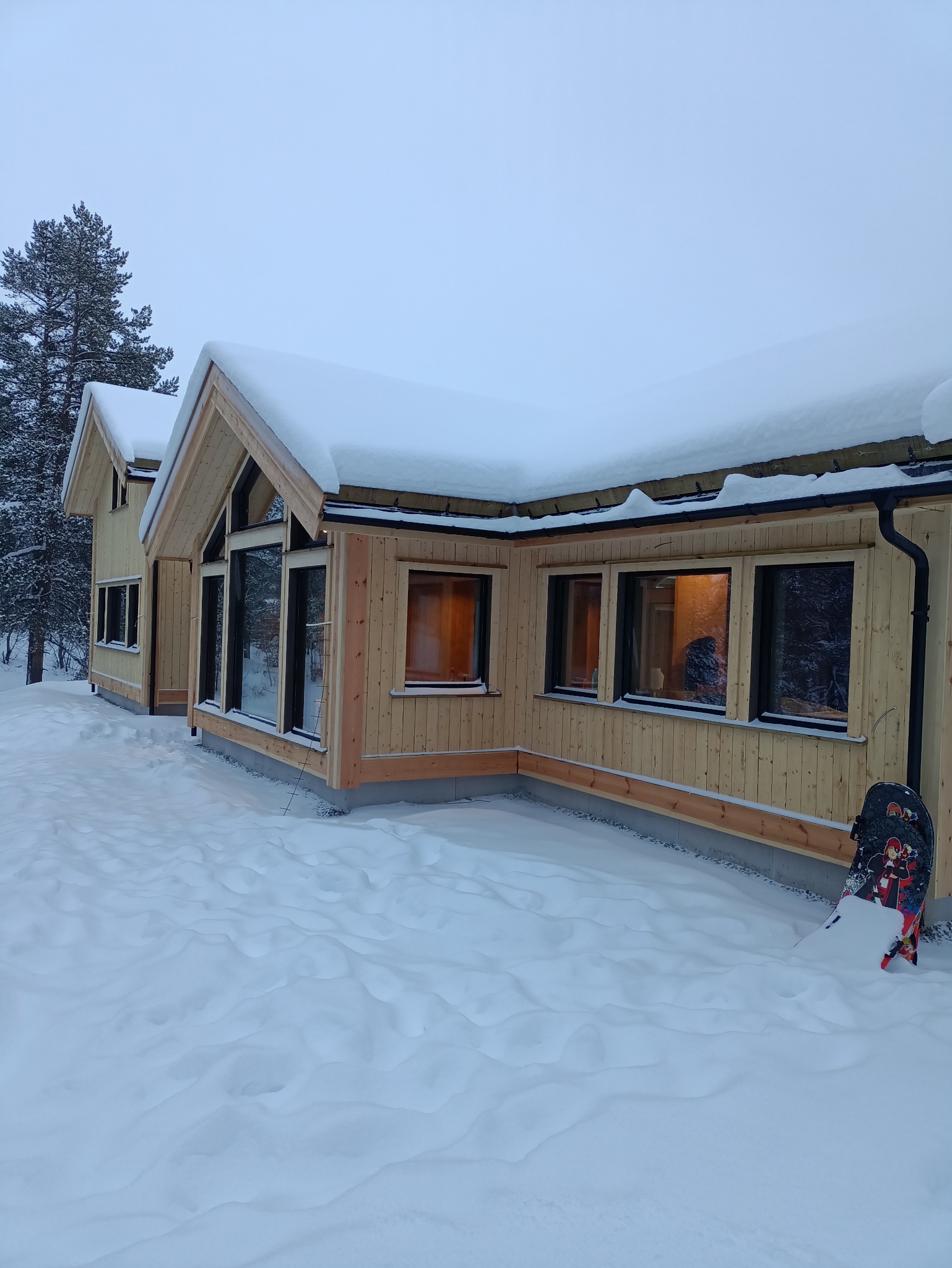 Surowy drewniany dom w zimowej scenerii, z dachem pokrytym śniegiem i snowboardem opartym o ścianę. Widoczne rynny i okna z ciepłym światłem wewnątrz.