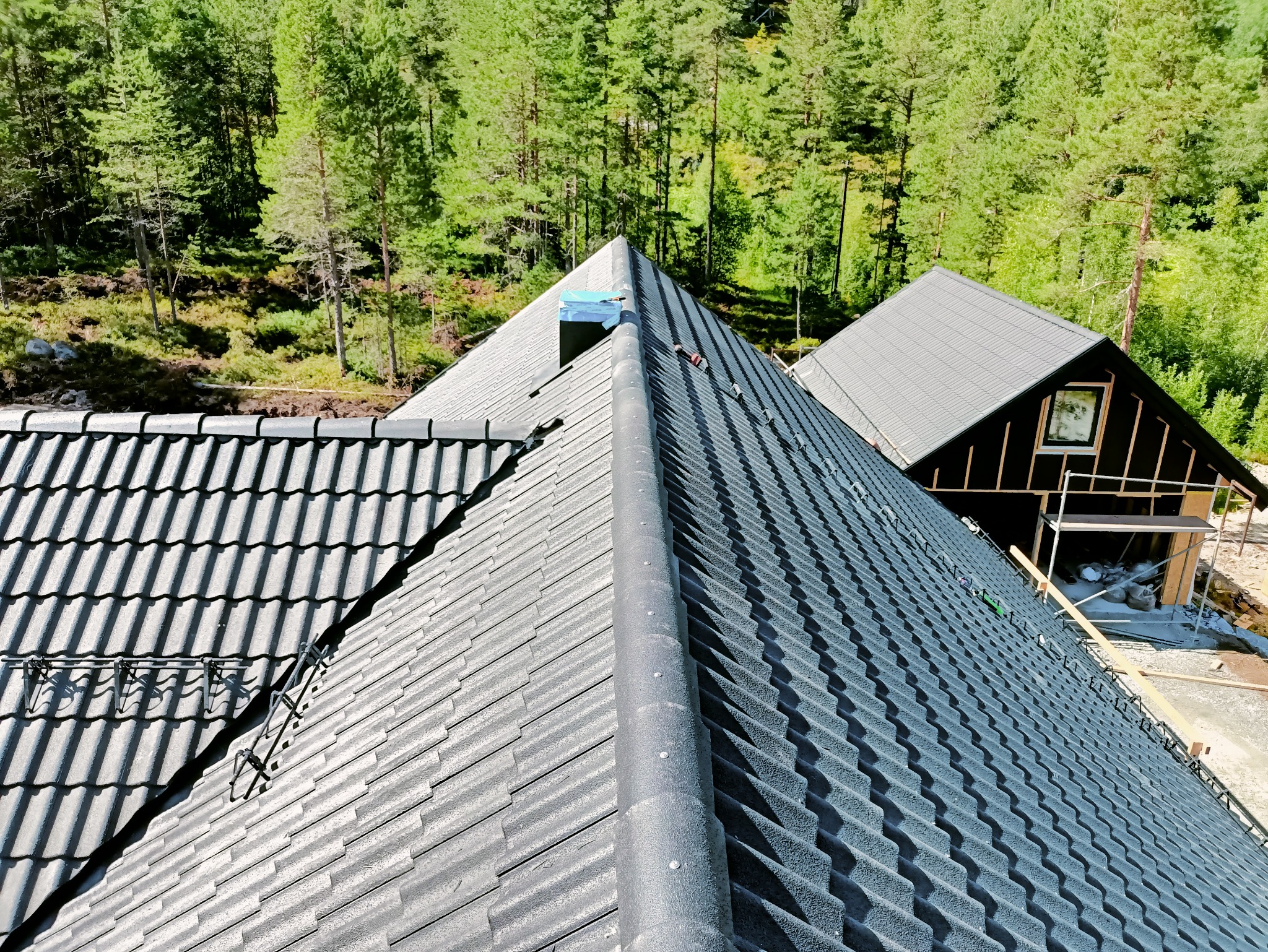 Szary dach z dachówki ceramicznej, widok z góry na tle zielonego lasu. Na dachu widoczne elementy montażowe i komin w trakcie instalacji. Obok budynek w stanie surowym.