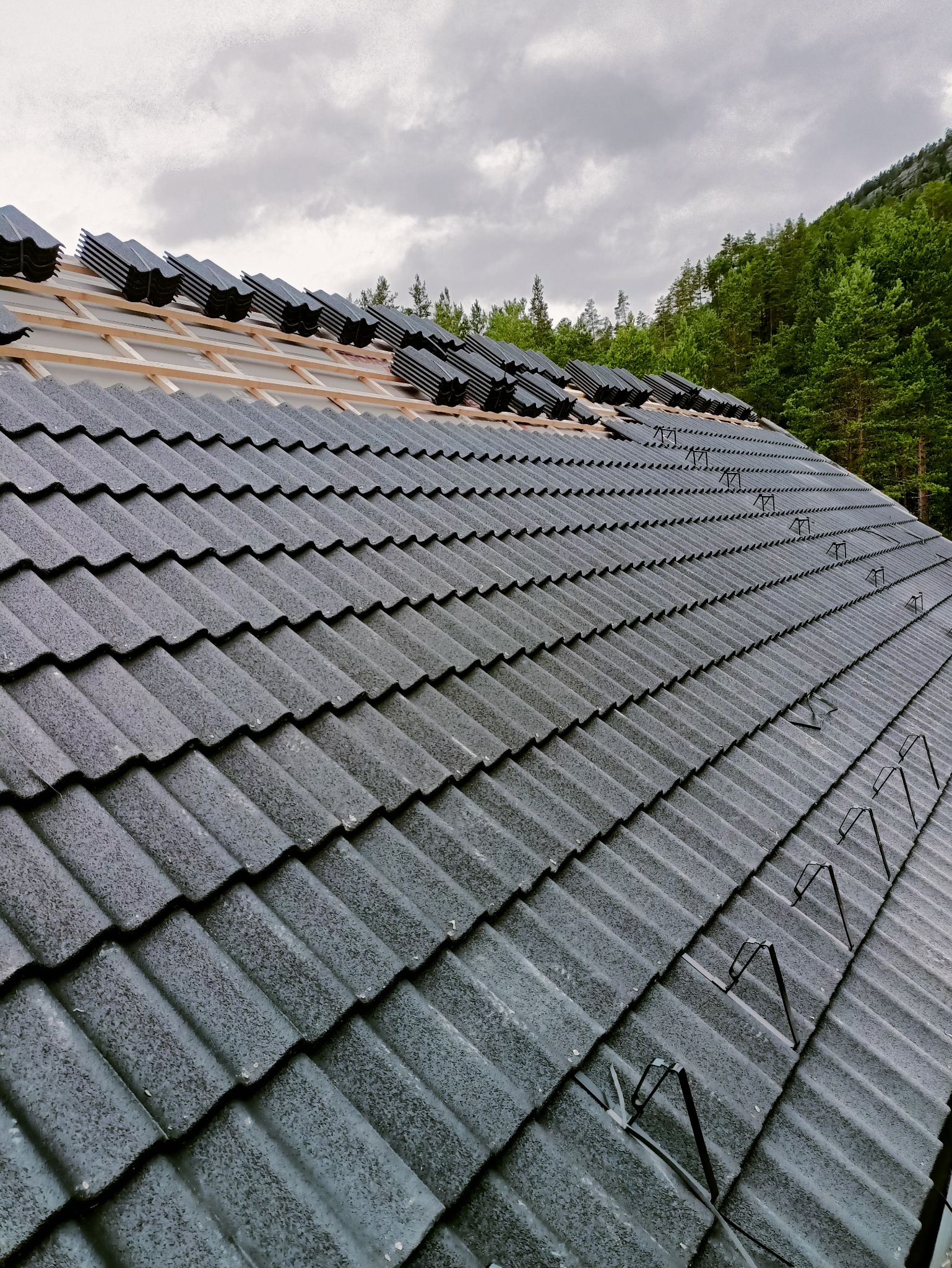 Szary dach z dachówki ceramicznej w trakcie układania, widoczna więźba i fragment lasu w tle. Częściowo ułożone dachówki na drewnianej konstrukcji.