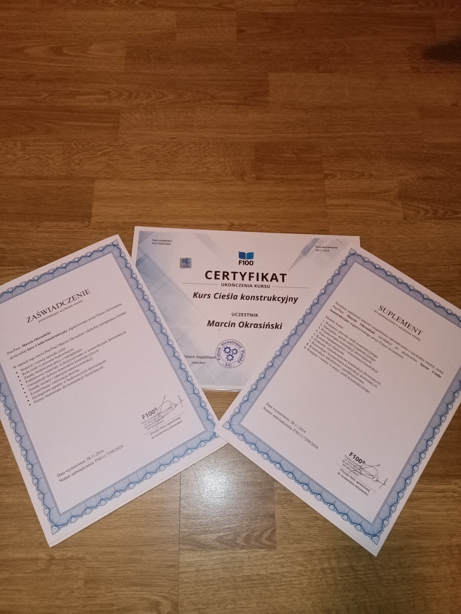 Certyfikat ukończenia kursu ciesielstwa konstrukcyjnego Marcin Okrasiński, suplement i zaświadczenie na drewnianym tle. Dokumenty z datą 28.11.2024.
