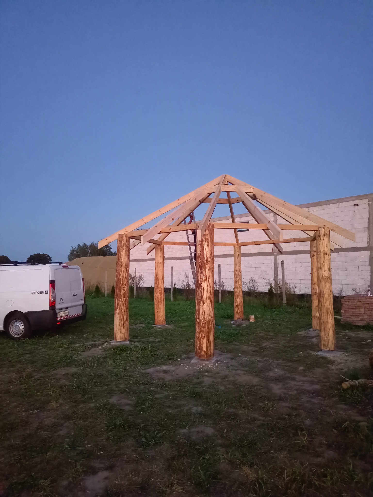 Wczesny etap budowy drewnianej altany z bali, widoczna konstrukcja dachu i słupy, na trawiastym terenie, z białym busem w tle i budynkiem w trakcie budowy.