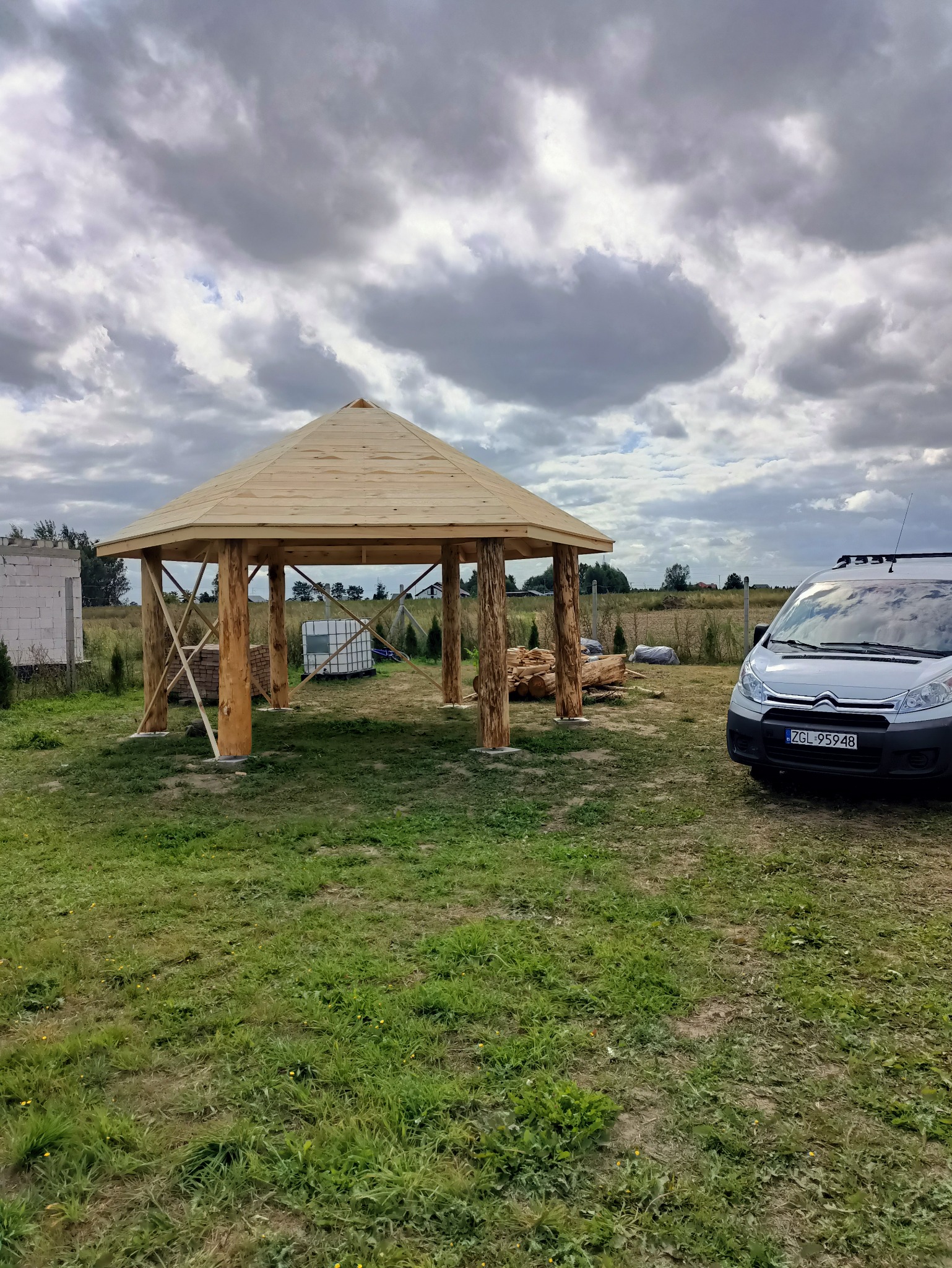 Surowa drewniana altana z bali na trawiastym terenie, częściowo ukończona, z widocznym stosem drewna i samochodem dostawczym w tle, pod pochmurnym niebem.