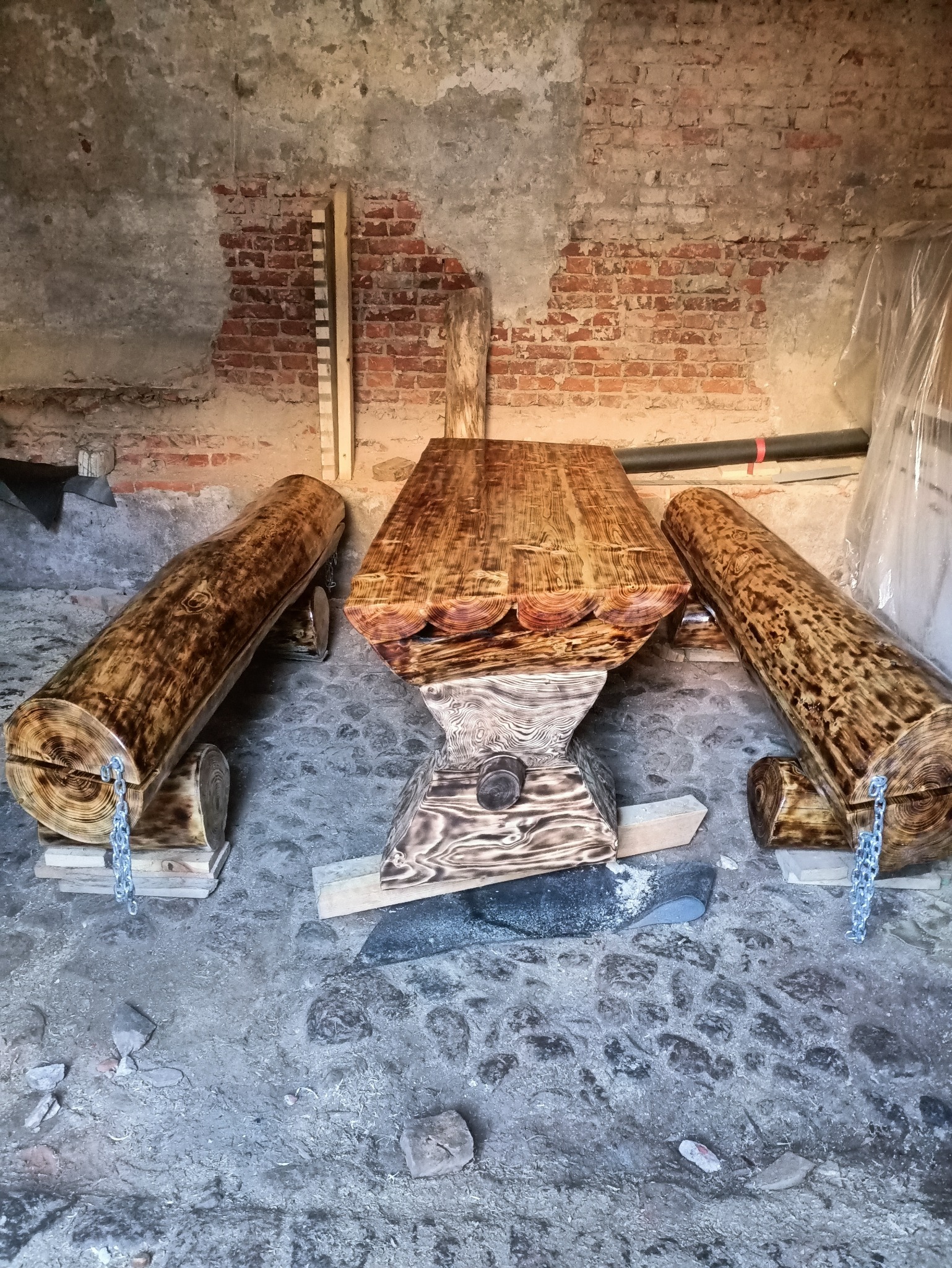 Rustykalny stół z drewna i dwa pufy z bali, połączone łańcuchem, na tle ceglanej ściany. Unikalne meble z naturalnych materiałów.