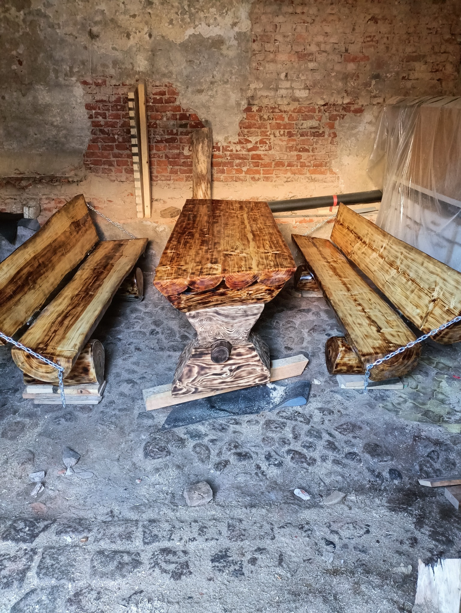 Rustykalny stół z dwoma ławkami z surowego drewna, połączonymi łańcuchami, na tle ceglanej ściany. Unikalny design i wykończenie.