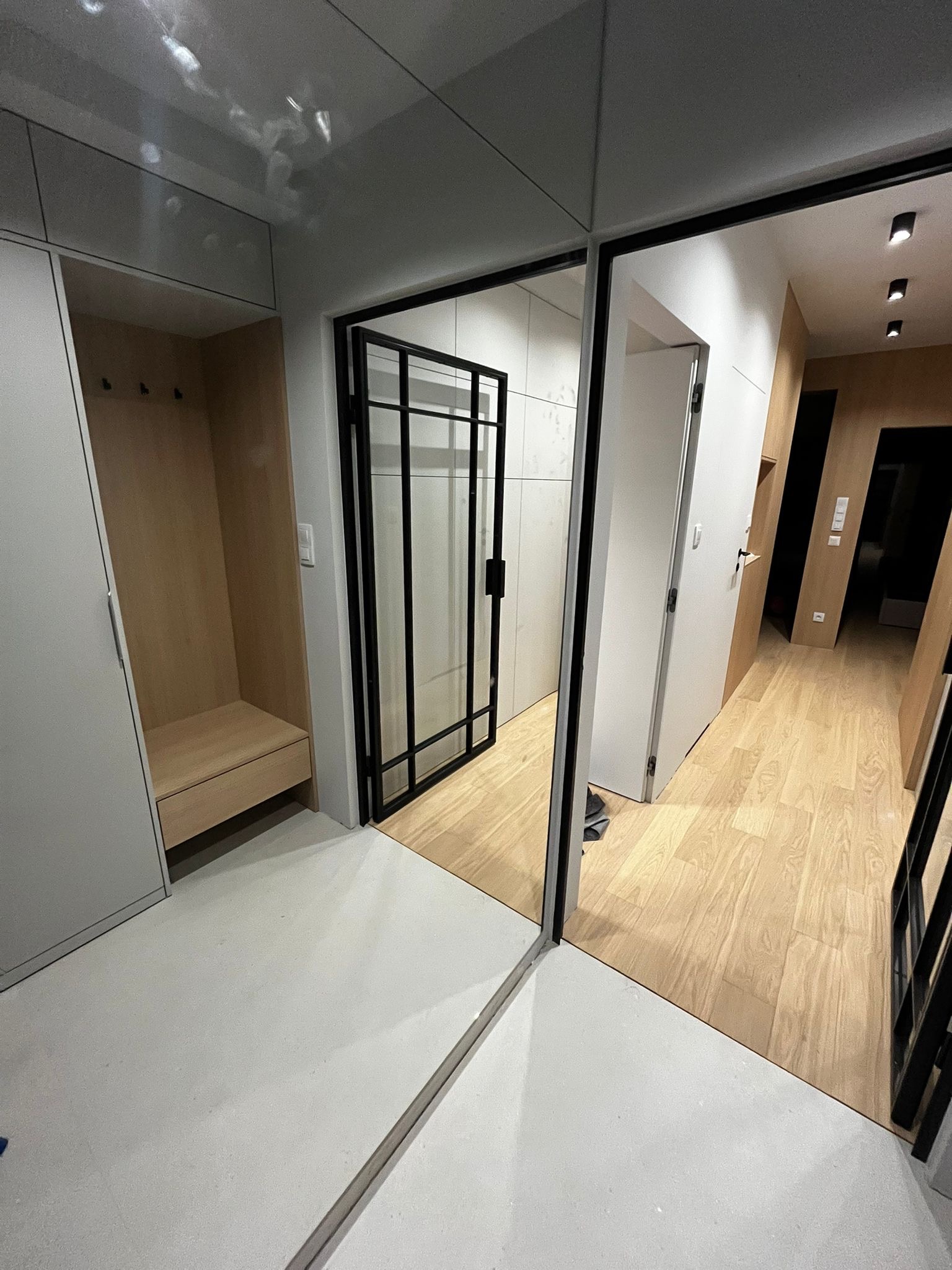 Nowoczesny przedpokój z szafą w zabudowie, drewnianą podłogą i czarną, przeszkloną ścianką działową. Minimalistyczny design, jasne kolory, oświetlenie punktowe.