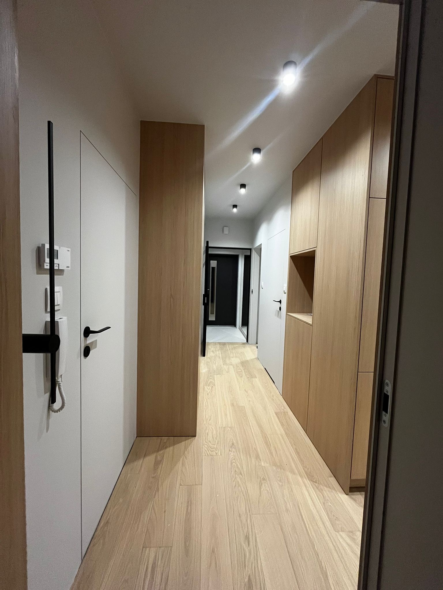 Nowoczesny korytarz z drewnianą podłogą, zabudową meblową z jasnego drewna i białymi drzwiami. Minimalistyczne oświetlenie sufitowe.