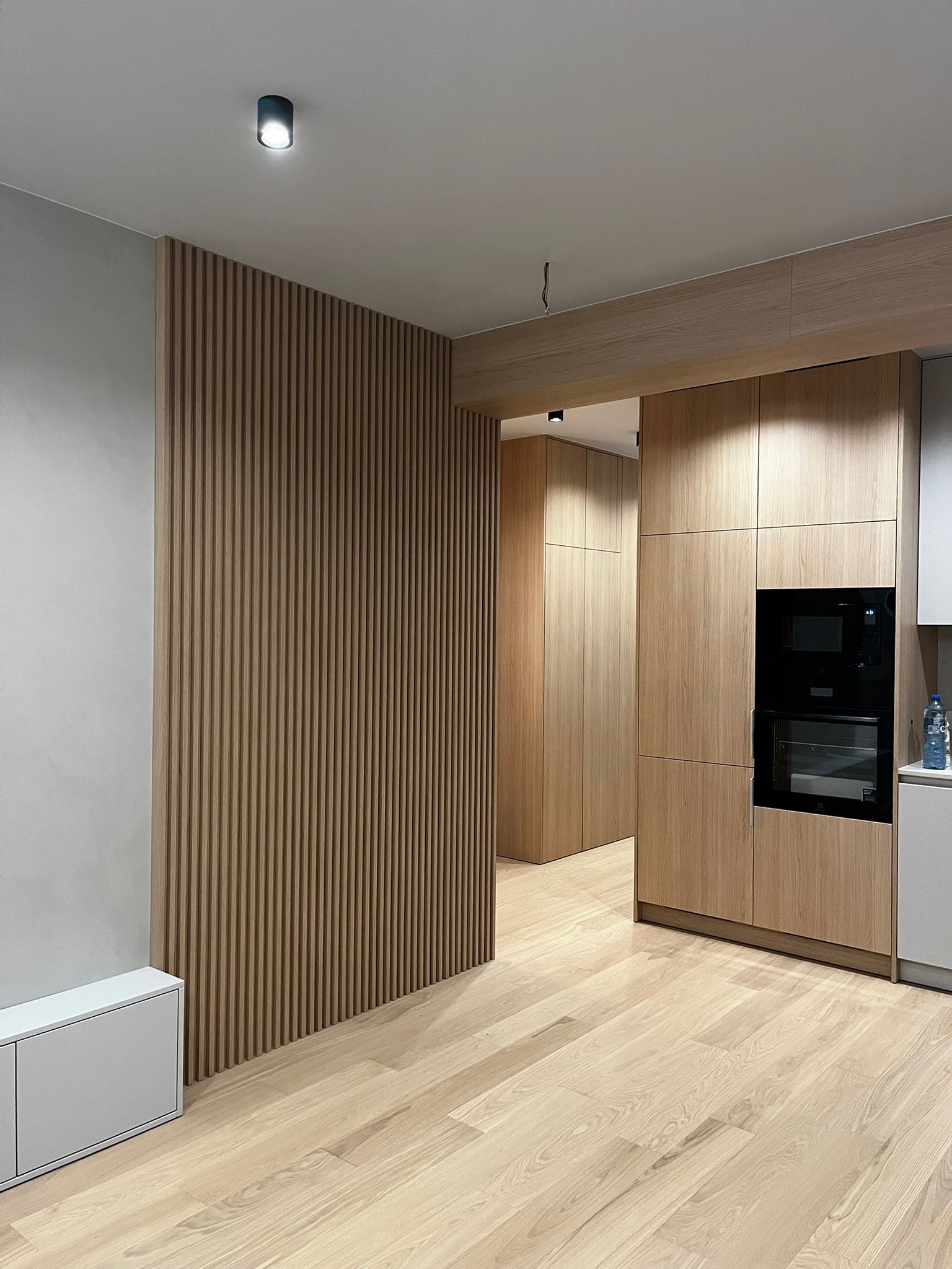 Nowoczesne wnętrze: drewniana lamelowa ściana, zabudowa kuchenna z piekarnikiem i jasna podłoga z desek. Minimalistyczny design z subtelnym oświetleniem.