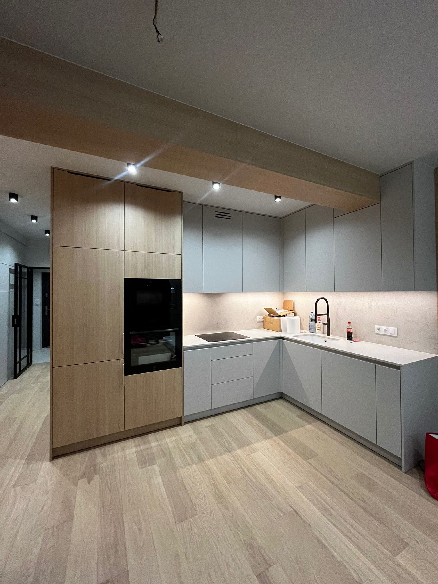 Nowoczesna kuchnia w odcieniach szarości i drewna z oświetleniem punktowym, widok na drewnianą podłogę i fragment otwartego przejścia do innego pomieszczenia.