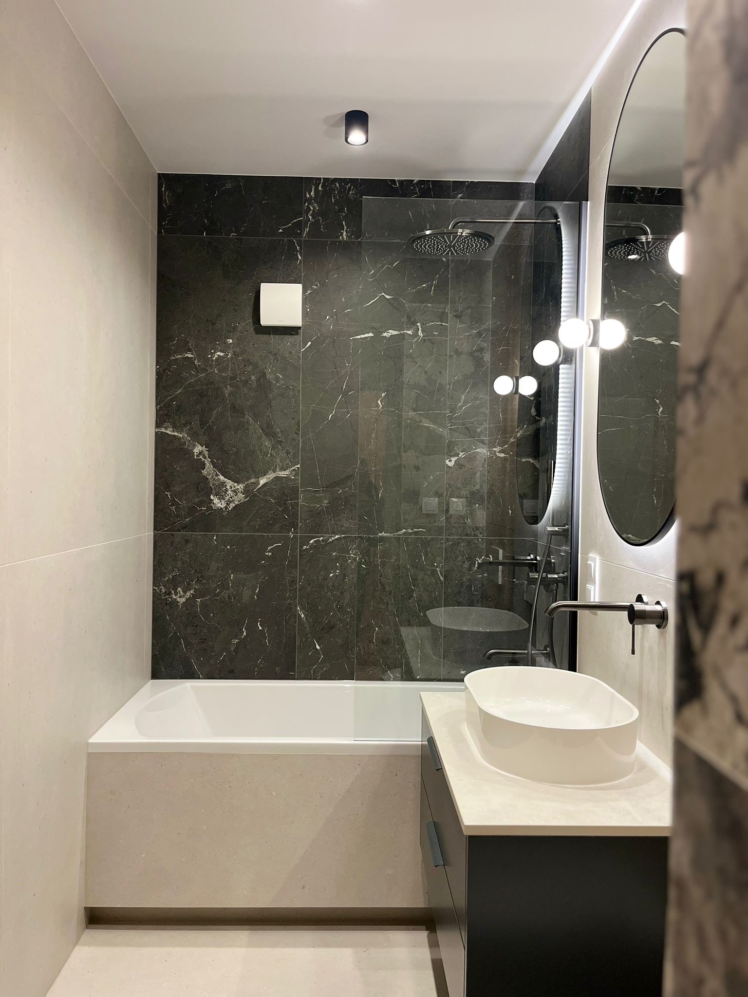 Nowoczesna łazienka z wanną obudowaną beżowymi płytkami, czarną ścianą z marmuru, owalnym lustrem z podświetleniem i białą umywalką nablatową.