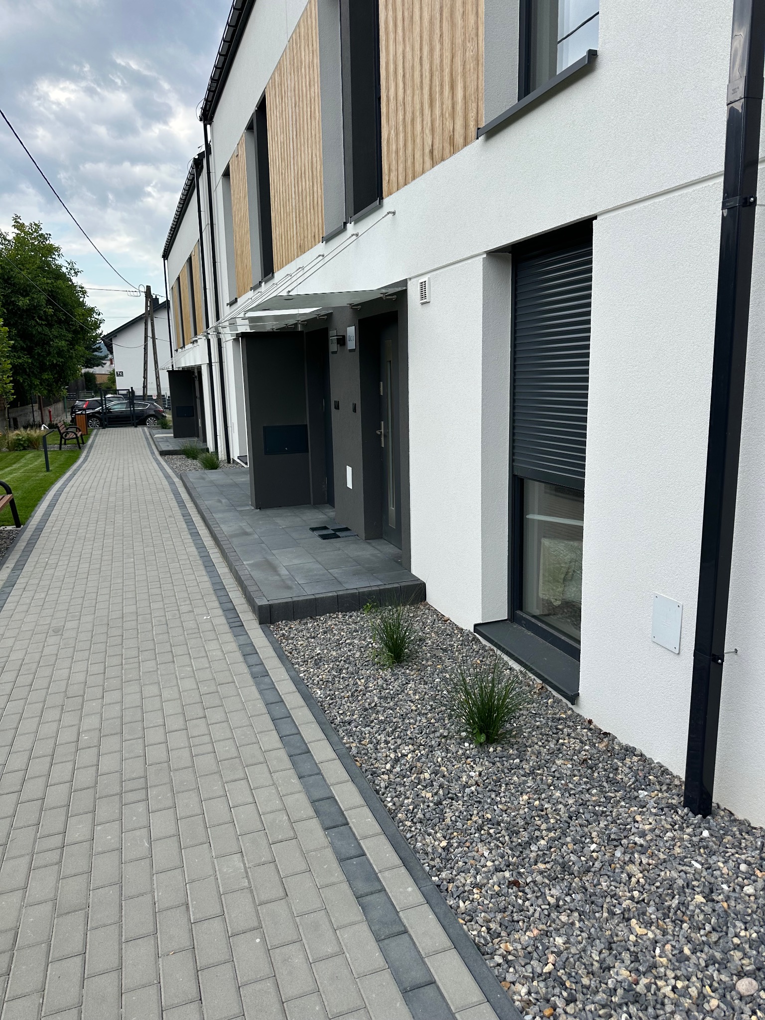 Nowoczesne szeregówki z elewacją w bieli i drewnie, szare okna z roletami, chodnik z kostki brukowej i kamienny ogródek. Minimalistyczny design.