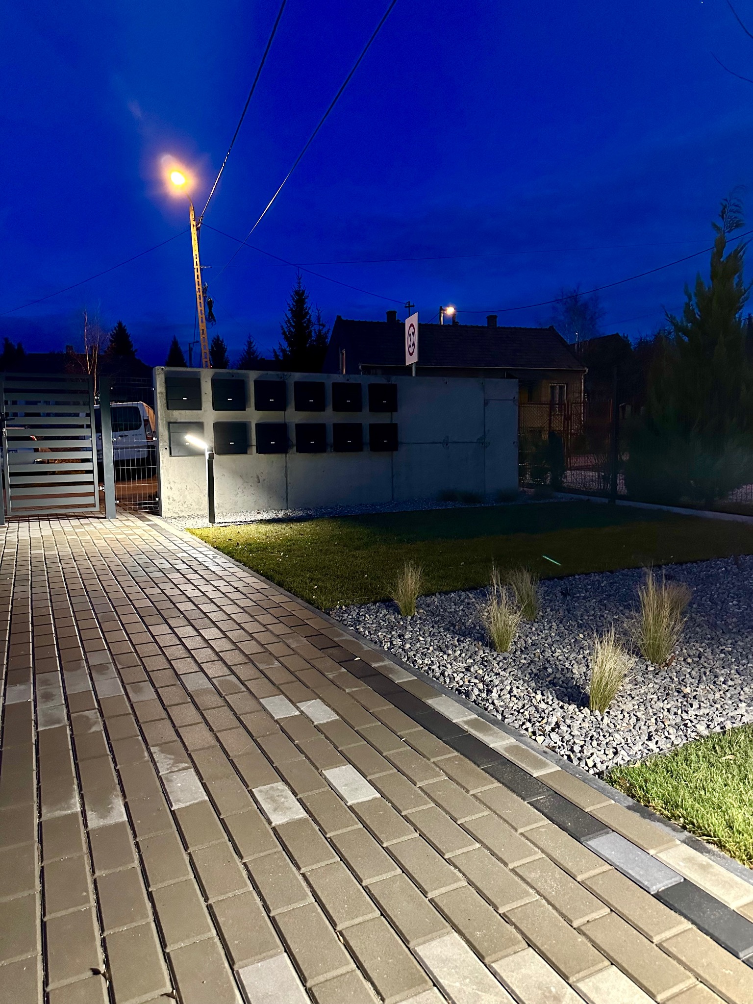 Oświetlona, nowoczesna posesja z brukowanym podjazdem, minimalistycznym ogrodzeniem i skrzynkami pocztowymi w betonowej ścianie, sfotografowana o zmierzchu.