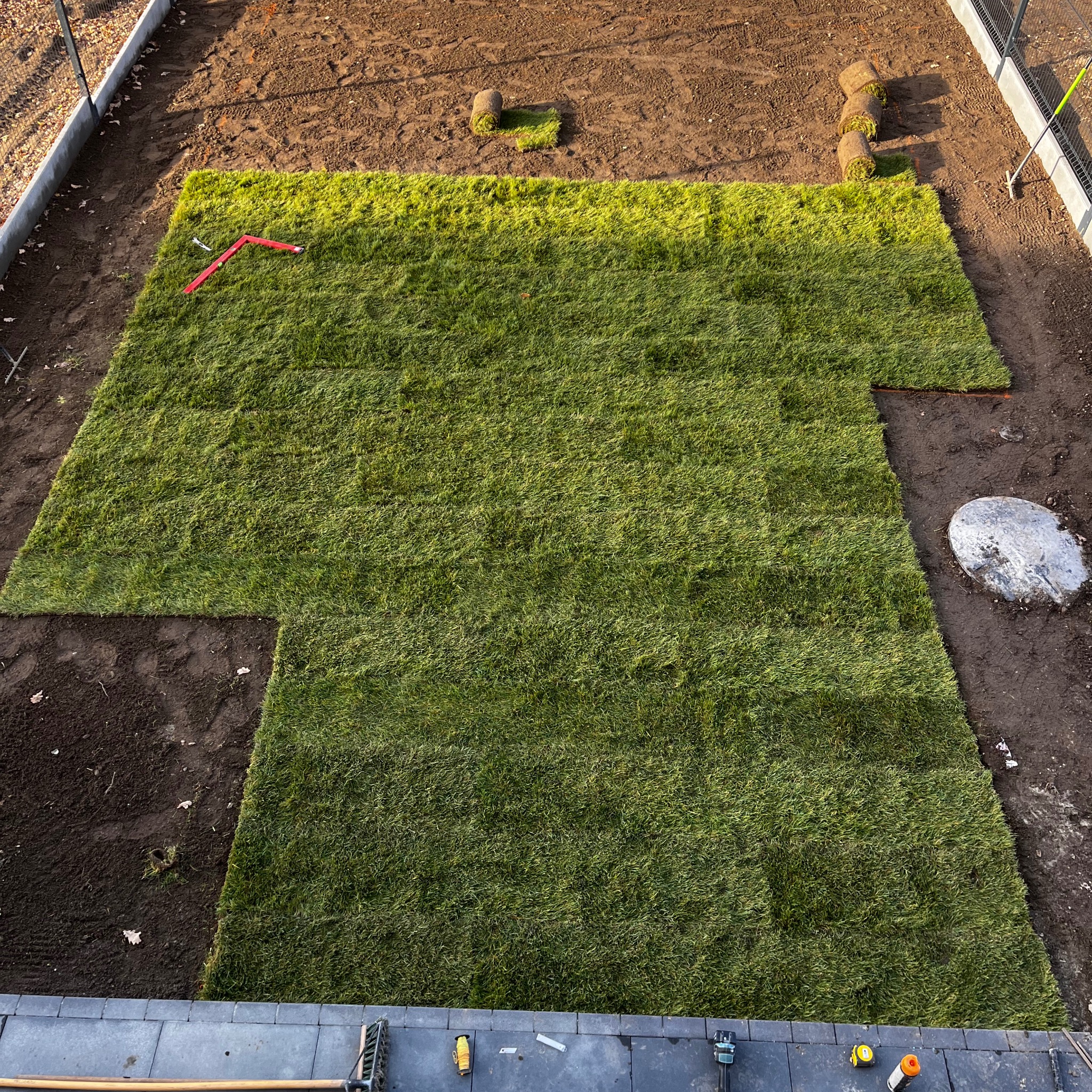 Układanie trawy z rolki na działce, widok z góry. Częściowo ułożona trawa, narzędzia ogrodnicze i rolki trawy w tle. Etap prac w słoneczny dzień.