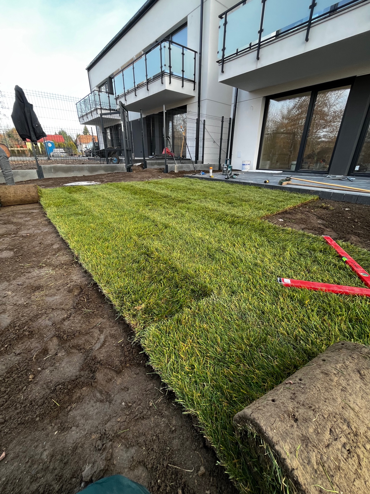 Układanie trawy z rolki przed nowoczesnym domem. Częściowo ułożony trawnik, widoczne narzędzia i rolka trawy. Oświetlenie naturalne, pochmurne niebo.